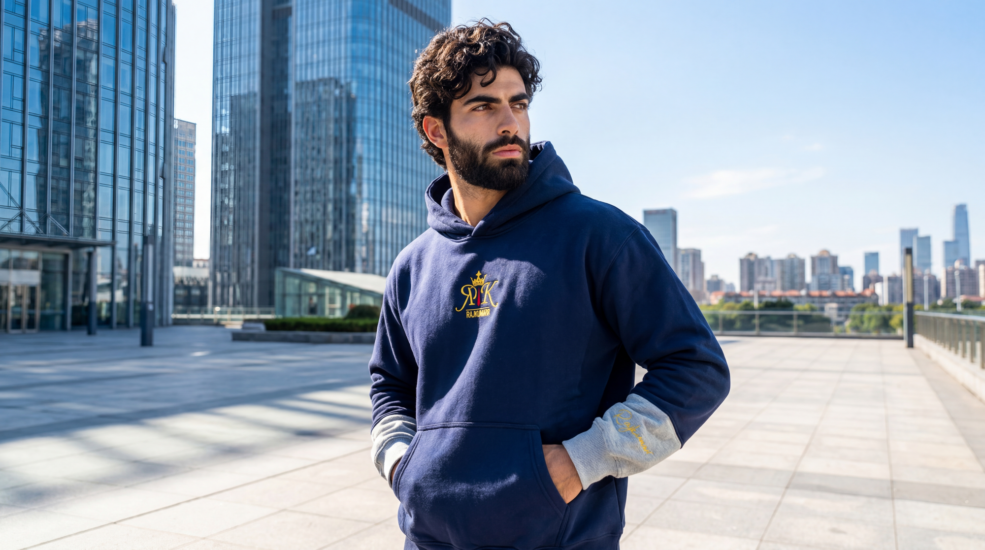 Model wearing a navy blue hoodie with a logo, standing in an urban setting with modern buildings.